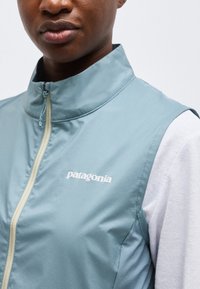 Άτομο που φορεί γαλάζιο αμάνικο γιλέκο Patagonia με φερμουάρ πάνω από ένα λευκό μακρυμάνικο πουκάμισο, δείχνοντας το πάνω μέρος του στήθους και της περιοχής του γιακά.