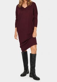 Robe bordeaux à coupe décontractée avec un col en V et des manches trois-quarts, associée à des bottes en cuir noires montantes jusqu'au genou.