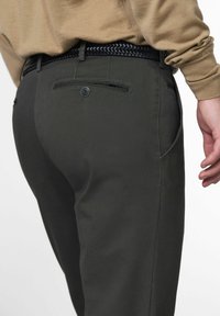 Close-up van een persoon met een donkere grijze gestructureerde broek, een zwarte geweven riem en een beige langwerpig shirt dat erin is gestoken.