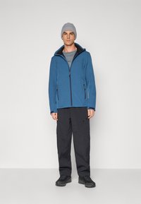 Blue softshell jacket with a zip front and hood, worn over a gray shirt, paired with black cargo pants and black hiking boots.