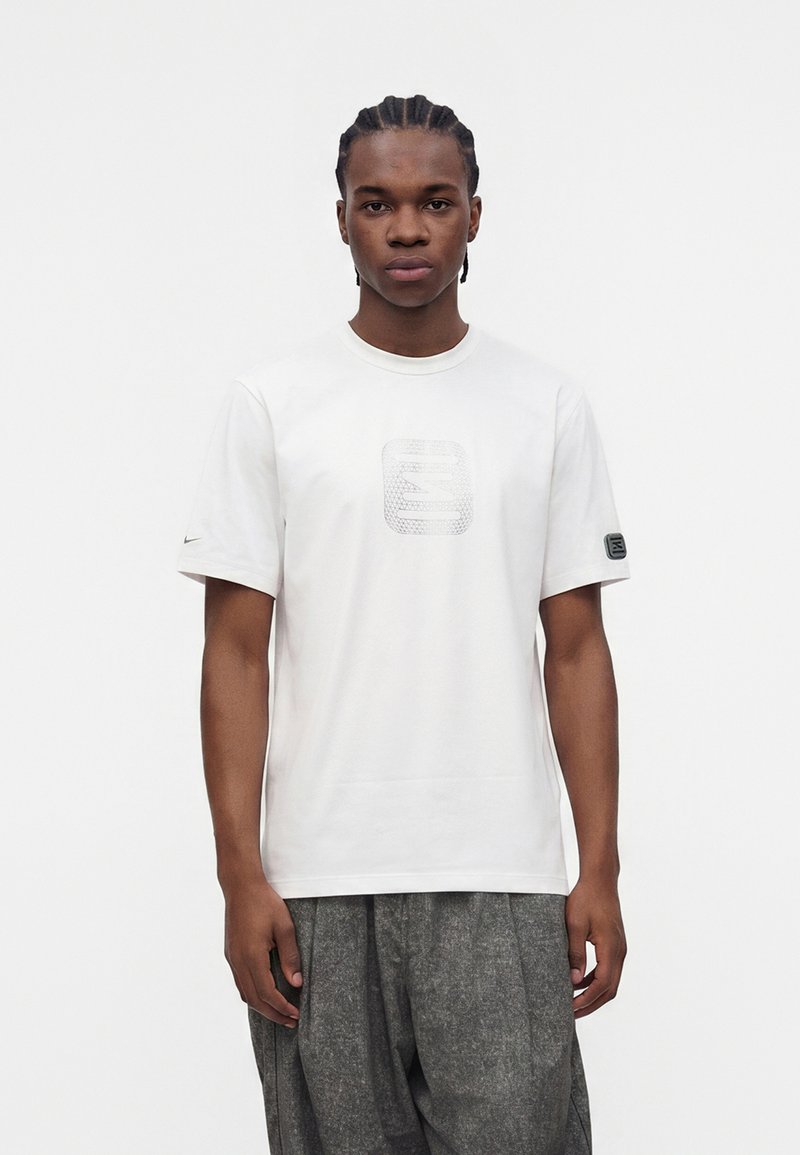 Young man with braided hair wearing white short-sleeve T-shirt with patterned square logo and loose gray pants, standing against white background.