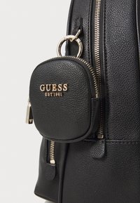 Svart lærryggsekk med en rund glidelåsveske med gull "GUESS"-logo. Synlige gullglidelåser og teksturert overflate.
