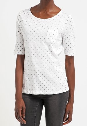Personne à la peau foncée portant une chemise blanche à manches courtes avec de petits pois noirs et une poche poitrine, associée à un pantalon gris foncé.