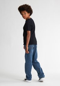T-shirt nera in cotone con maniche corte, abbinata a jeans in denim blu a vestibilità ampia e sneakers bianche. I capelli sono acconciati in un afro riccio.