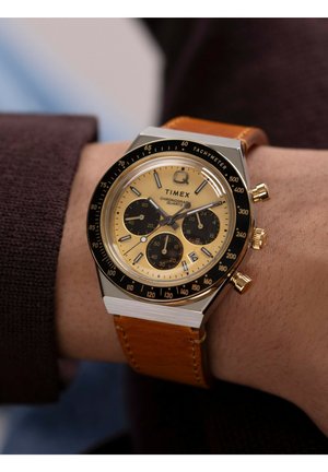 La montre chronographe Timex présente un cadran doré, des sous-cadrans noirs, un boîtier en acier inoxydable et un bracelet en cuir beige. Lunette tachymétrique incluse.