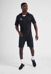 Atletisk man i svart sportklädsel och sneakers, stående med en lätt rörelse mot en enfärgad ljus bakgrund.