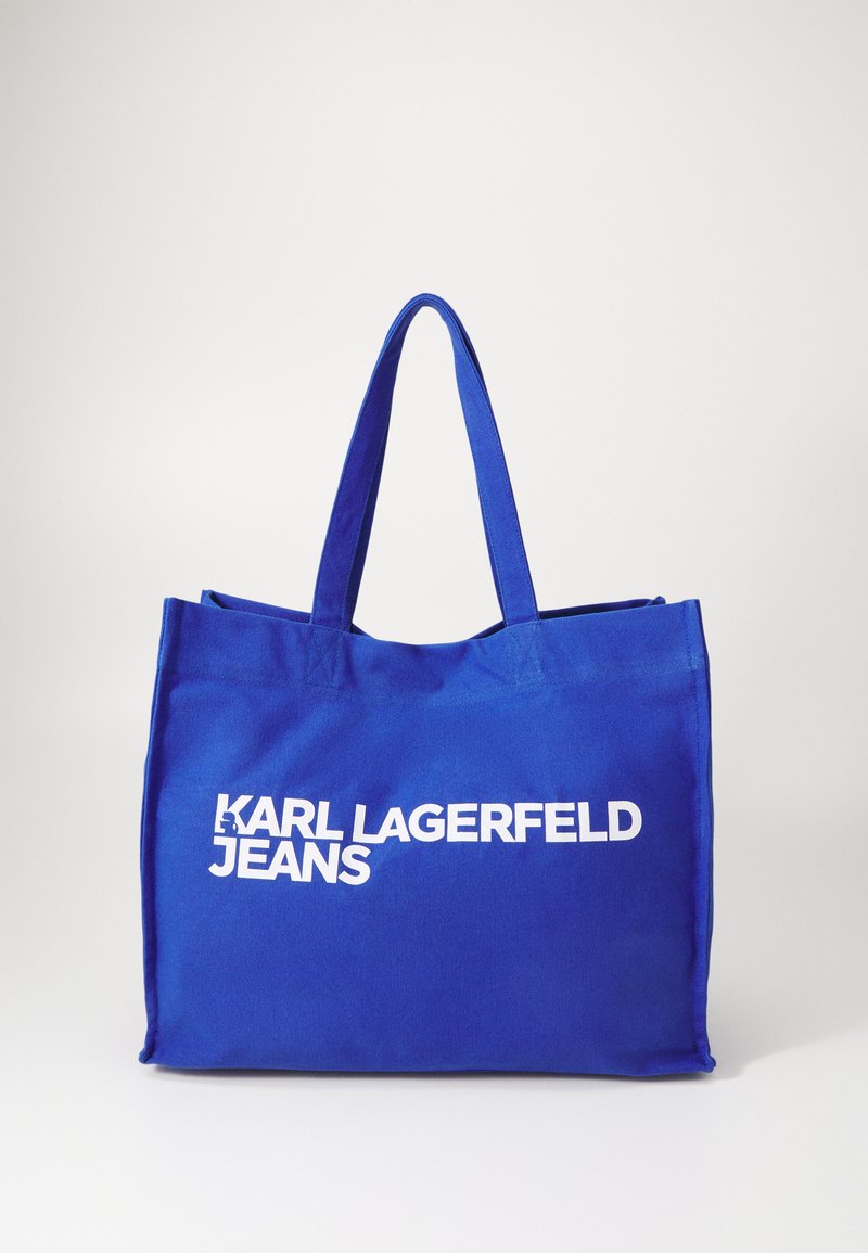 Mėlynas kanvos pirkinių krepšys su dviem rankenomis, su dideliu baltu tekstu, kuris skelbia "KARL LAGERFELD JEANS" priekyje. Paprastas dizainas.