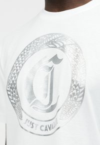 Λευκό T-shirt με μεταλλικό ασημί λογότυπο που παρουσιάζει ένα στυλιζαρισμένο γράμμα "J" περιτριγυρισμένο από ένα σχεδιασμό ελικοειδούς φιδιού και το κείμενο "Just Cavalli".