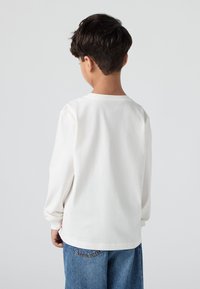 Bijela pamučna majica dugih rukava s okruglim izrezom, prikazana s leđa, u kombinaciji s plavim trapericama. Tkanina izgleda mekano i glatko.