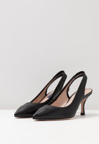 Des escarpins en cuir noir à dos ouvert avec des bouts pointus et des talons aiguilles de hauteur moyenne, présentés sur une surface blanche contre un fond uni.