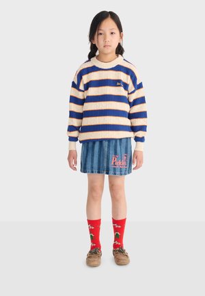 Dievča s tmavými vlasmi vo vrkočoch, oblečená v modro-béžovom pruhovanom svetri, rifľovej sukni s ružovým nápisom, červených ponožkách so vzorom a hnedej obuvi typu loafers.