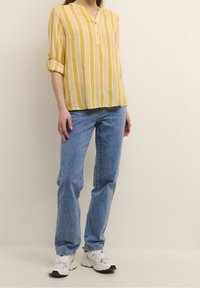Gulrandig blus med V-ringning och knappdetalj, kombinerad med ljusblå jeans och vita sneakers. Blusen har uppvikta ärmar.