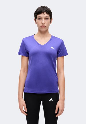 Persoană tânără cu păr scurt și închis la culoare, purtând un tricou mov cu guler în V și pantaloni negri, ambele cu logo-uri albe Adidas, stând drept și privind înainte.