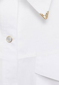 Chemise blanche avec des accents décoratifs dorés sur le col, un bouton rond blanc avec un bord doré, et une texture de tissu lisse.