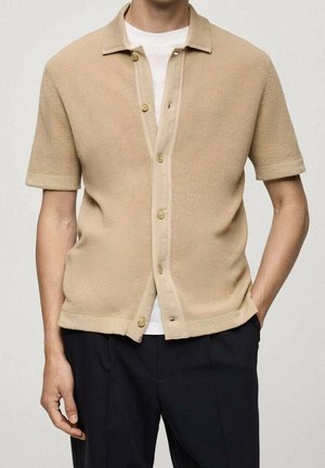Homme portant une chemise en maille beige à manches courtes et boutons par-dessus un T-shirt blanc, avec un pantalon noir, les mains reposant le long du corps.