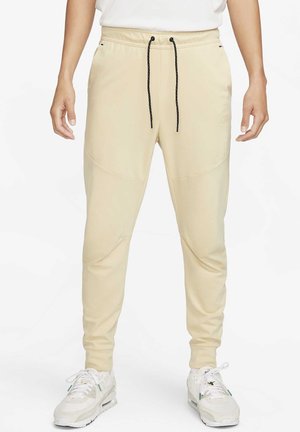 Pantalon de survêtement beige fuselé avec cordons noirs porté par une personne en baskets blanches et haut blanc, debout les bras détendus le long du corps.