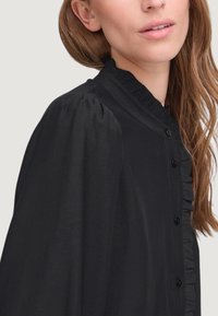 Blusa nera con colletto a volant e placca, dotata di maniche a sbuffo e piccoli bottoni neri lungo la parte anteriore. Texture liscia.