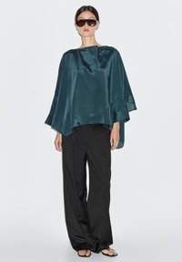 Teal satin poncho med et draperet design, parret med løstsiddende sorte bukser og sorte sandaler, båret med oversized solbriller.
