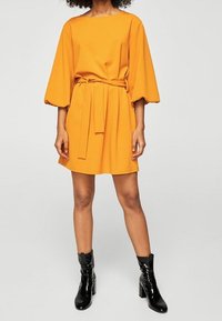 Krótkoskrzydłowa, żółta sukienka z paskiem w talii, o luźnym kroju i teksturowanej tkaninie, w połączeniu z błyszczącymi czarnymi botkami do kostek.