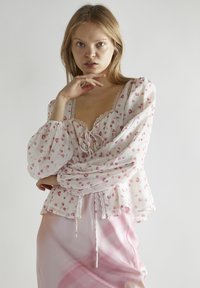 Blusa bianca floreale con accenti rosa, maniche a sbuffo, rifiniture in pizzo e dettaglio a fiocco sul davanti. Indossata sopra una gonna con sfumature di rosa.