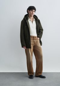 Σκούρο πράσινο μπουφάν με κουκούλα πάνω από μια κρεμ ζακέτα με φερμουάρ, σε συνδυασμό με ανοιχτό καφέ παντελόνι φαρδιάς γραμμής και μαύρα παπούτσια, πάνω σε ουδέτερο φόντο.