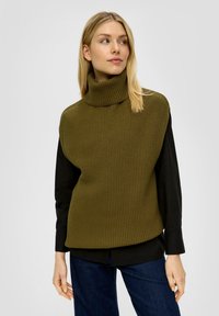 Khaki, geribbelde coltrui over een zwart langslavend shirt, gecombineerd met donkerblauwe jeans. Soepele stof, relaxte pasvorm.