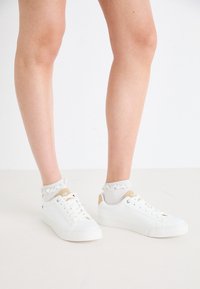 White sneakers with a smooth texture and minimalistic design. Accented with a beige heel tab and worn with light gray frilled socks.