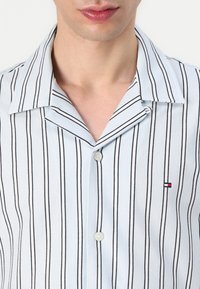 Camisa de manga corta azul claro con rayas verticales negras, cuello con muesca y cierre de botones. Pequeño logotipo en el lado izquierdo.
