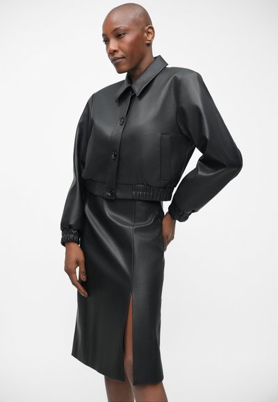 Crna lažna kožna kratka jakna s kopčanjem na gumbe i elastičnim naramenicama, uparena s uskom crnom kožnom suknjom s bočnim prorezom.
