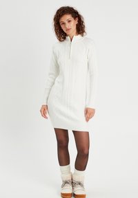 Robe en tricot blanc avec un col haut et une fermeture éclair, arborant un motif en tricot torsadé et un bas en côtes. Le modèle porte des collants noirs et des bottes en fourrure.