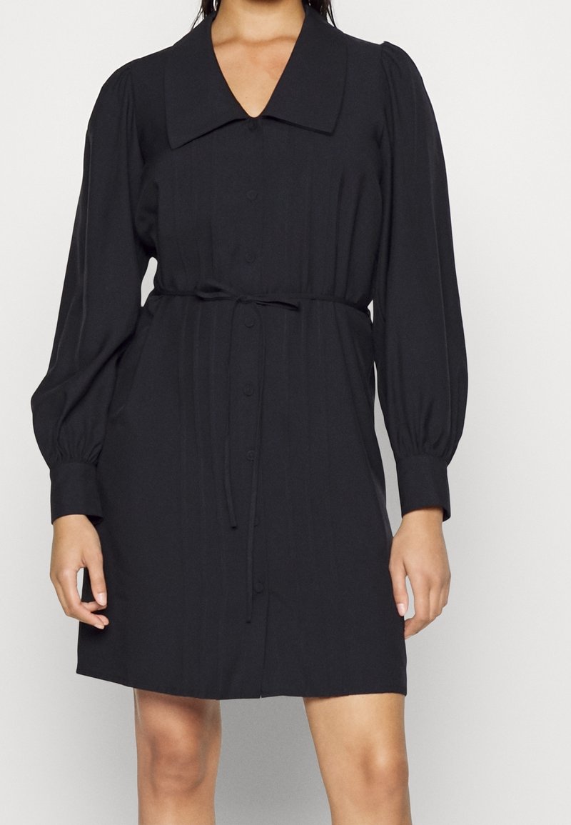 Femme portant une robe noire boutonnée avec un col large, des manches longues bouffantes et une ceinture nouée à la taille.