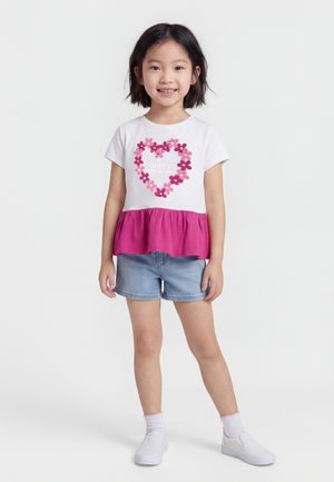 Młoda dziewczyna stoi, ubrana w białą i różową koszulkę z kwiatowym sercem i napisem "Guess Love Story", dżinsowe szorty, białe trampki, uśmiecha się.