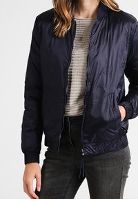Marineblaue Daunenjacke mit rundem Kragen, Reißverschluss und gerippten Strickbündchen. Getragen über einem hellgrauen strukturierten Pullover und dunklen Jeans.