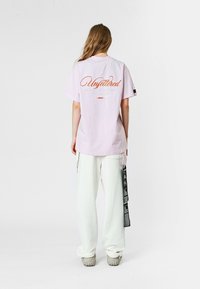 Camiseta de algodón rosa claro con "Sin ataduras" bordado en naranja en la parte trasera, combinada con pantalones blancos que presentan un detalle de etiqueta negra.