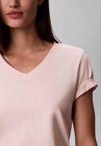 Γυναίκα που φοράει ανοιχτοροζ T-shirt με V λαιμόκοψη, γυρισμένα μανίκια και μικρό λευκό λογότυπο "cK" στο αριστερό στήθος, σε γκρι φόντο.