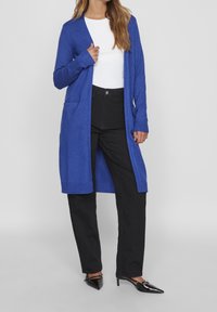 Cárdigan de punto azul con mangas largas y abertura frontal, con puños acanalados y bolsillos, combinado con pantalones negros y tacones negros.