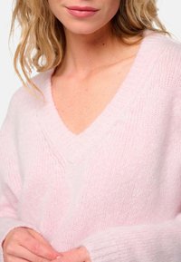 Pull en tricot rose clair avec un col en V, des poignets côtelés et une texture douce, affichant une coupe décontractée et des détails minimalistes.