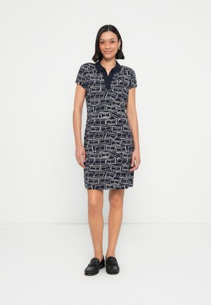 Kvinde iført en navyblå polo kjole med korte ærmer med hvide abstrakte linjemønstre og sorte loafers, stående foran en ensfarvet hvid baggrund.