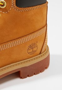 Stivale in suede marrone con colletto imbottito nero, occhielli dorati, cuciture bianche e suola in gomma marrone con impressa un logo ad albero.