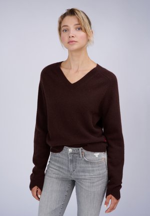 Blonde vrouw met een donkerbruine V-hals trui en lichtgrijze spijkerbroek, staand tegen een effen lichte achtergrond.
