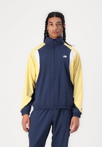 Veste de survêtement navy et jaune avec des accents blancs, zip devant, col montant, poignets et taille élastiques, conçue pour un ajustement sportif.