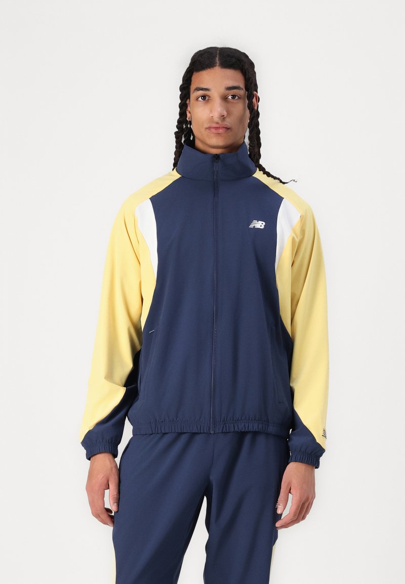 Veste de survêtement navy et jaune avec des accents blancs, zip devant, col montant, poignets et taille élastiques, conçue pour un ajustement sportif.