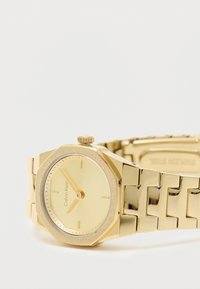 Zelta metāla pulkstenis ar astoņstūrainu korpusu, gludu dzeltenu ciparnīcu, minimālistiskiem marķieriem un masīvu aproces dizainu ar spīdīgiem posmiem.