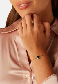 Bracelet en or avec une gemme verte ovale entourée de petites pierres claires, portée sur une main contre une chemise en satin rose.