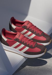 Paar rote und braune Adidas-Sneaker mit weißen Streifen und Gummisohlen, die auf einer weißen Oberfläche mit Schatten liegen.