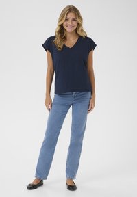 Top azul marino con cuello en V y mangas cortas, combinado con jeans de pierna recta de color azul claro y zapatos planos negros, frente a un fondo blanco liso.