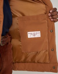 Bruine geïsoleerde jas met een gladde satijnen voering, een zak met inzetstuk en een label met een citaat. Drukknoopsluiting langs de zoom.