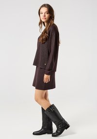 Burgunderfarbener Strickpullover mit glitzernder Textur, locker sitzend über einem passenden Rock. Das Model trägt knielange schwarze Lederstiefel mit Schnallen.