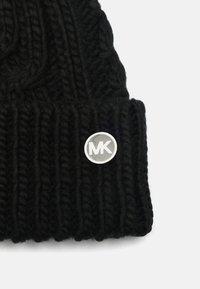 Gorro de punto negro con un patrón acanalado, hecho de hilo grueso, con un acento de logo circular plateado en el puño.