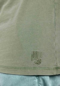 T-shirt en coton vert olive avec un tissu texturé et un logo brodé "PULL IN" en bas, agrémenté de détails de couture contrastants.